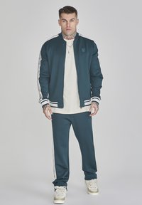Chándal deportivo en color verde azulado que incluye una chaqueta bomber con cremallera y rayas blancas, acompañada de pantalones a juego. Una camiseta interior blanca y zapatillas completan el conjunto.