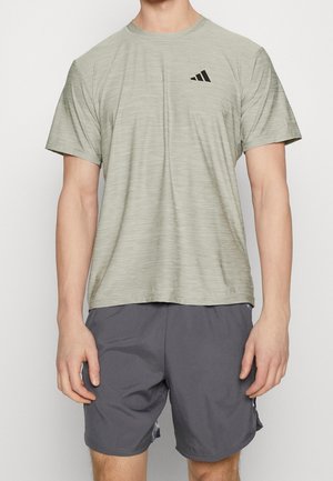 Mann trägt hellgraues Adidas T-Shirt mit kurzen Ärmeln und dunkelgraue Sportshorts, steht vor einem einfarbigen Hintergrund.