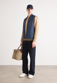 Colete sem mangas acolchoado na cor marinha por cima de um sweater bege, jeans escuros, tênis brancos e uma bolsa bege com detalhes e alças castanhas.