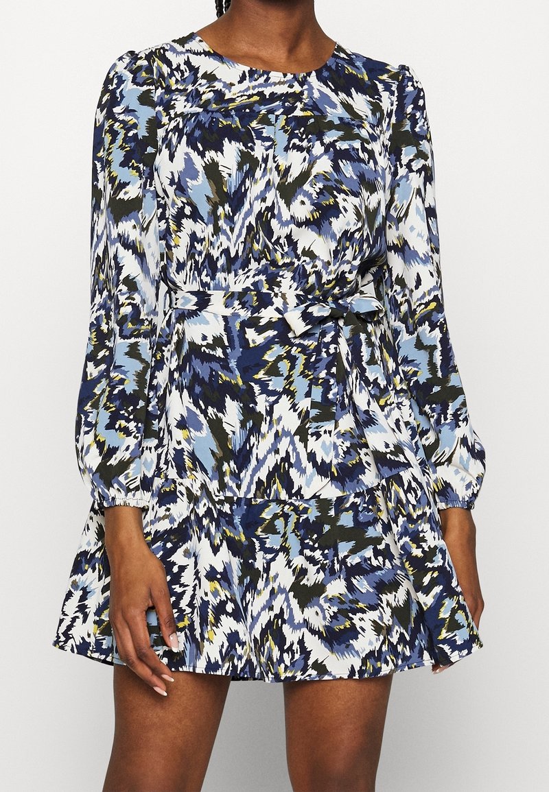Femme portant une robe à manches longues, ceinturée, avec un motif abstrait bleu, blanc et noir et un ourlet court à volants.