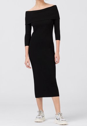 Vrouw in een zwarte off-shoulder midi-jurk met driekwart mouwen en witte chunky sneakers, staand tegen een effen achtergrond.