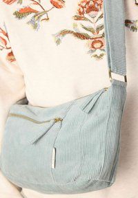 Sac bandoulière en velours côtelé bleu clair avec une fermeture éclair dorée, surface texturée et une poche avant supplémentaire. Bandoulière réglable attachée.