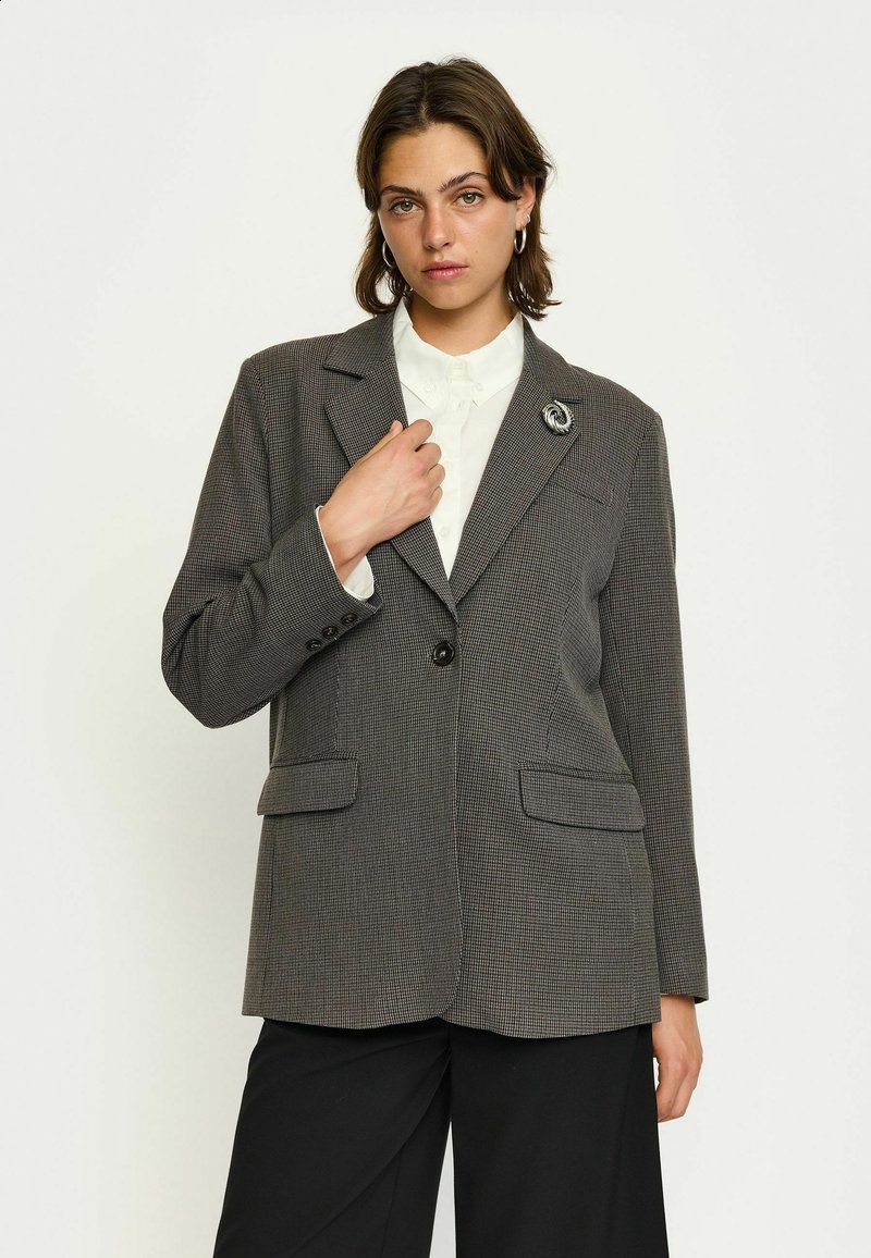 Blazer a quadri grigi con collo a scollatura, chiusura con un bottone, due tasche frontali e una spilla decorativa sulla parte sinistra del risvolto.