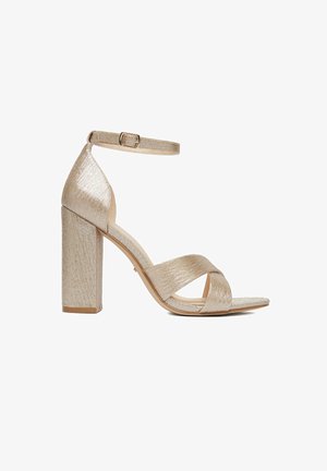 Sandalia de tacón alto con textura dorada, tacón ancho, tira en el tobillo con hebilla y tiras cruzadas sobre el dedo al descubierto.
