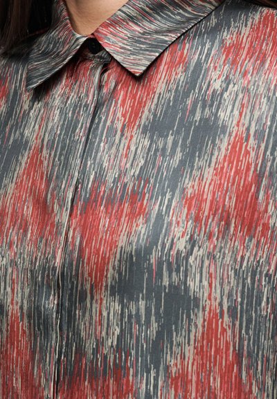 Krave og øverste del af brystet på en skjorte med rødt, gråt og hvidt abstrakt lodret stribemønster, knaplukket ved halsen.
