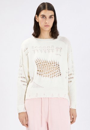 Donna dai capelli scuri che indossa un maglione bianco a maglia con grandi dettagli a maglia aperta e pantaloni rosa chiaro, in piedi davanti a uno sfondo semplice.