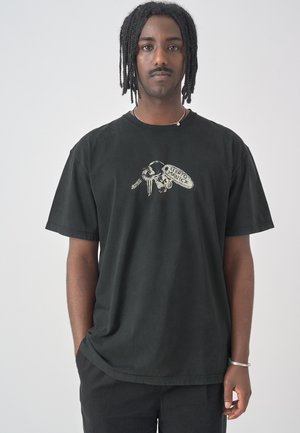 Junger Mann mit geflochtenem Haar, der ein schwarzes T-Shirt mit einem Schlüsselanhänger-Grafik und dem Text "Cierto Maniex," trägt, steht vor einem einfarbigen Hintergrund.