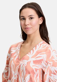 V-Ausschnitt-Bluse aus hellrosa Stoff mit einem weißen, organischen Blattmuster. Lässige Passform, weiche Textur und subtiler Glanz verleihen ihr ein besonderes Design.