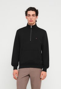 Μαύρο φούτερ με φερμουάρ τριών τετάρτων και ψηλό γιακά, με μπροστινή τσέπη και μικρή λεπτομέρεια λογότυπου στο στήθος. Μαλακό ύφασμα και ριμπ μανσέτες.