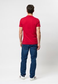 Tricou roșu cu mânecă scurtă expus din spate; fabricat din bumbac cu o textură netedă și o croială obișnuită; asortat cu blugi albaștri și adidași.
