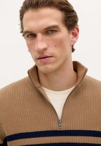 Pull en maille gaufrée, couleur camel, avec une fermeture éclair demi-zip et des rayures navy ; il possède une fermeture éclair en métal et un tissu léger. Design avec un col court.