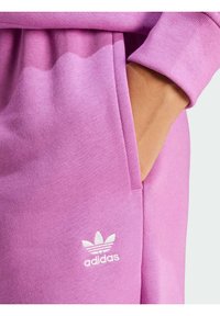 Rosa sweatpants med en slät textur, sidoficka och vit Adidas-logotyp med tre bladdesign nära nederkanten.