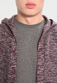 Sweat à capuche en maille texturée, bordeaux et grise, à ouverture frontale, porté sur un t-shirt gris uni. Comprend une capuche avec cordon de serrage et une coupe décontractée.