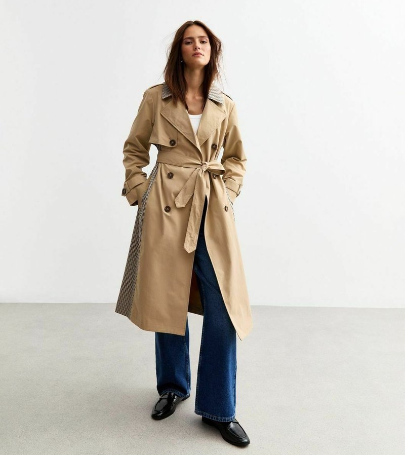 Beige trenchcoat med dubbelknäppta knappar, midja med bälte och rutiga krage accenter; kombinerad med vida blå jeans och svarta skor.