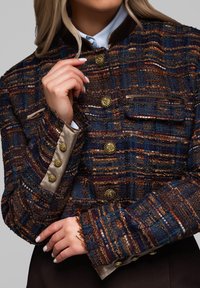 Marynarka tweedowa w odcieniach niebieskiego, pomarańczowego i brązowego z złotymi guzikami. Posiada wysoki kołnierz oraz teksturowane rękawy z przeszytymi detalami.