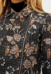 Blomster-mønstrede jakke i sort stof med metaltråde; har en lynlåslukning, krave og indviklet broderi i varme nuancer.