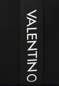 Fekete szövet táska, amelyen kiemelkedő fehér "VALENTINO" logó található, texturált anyaggal és lapos formával, strapabíró kialakítással.