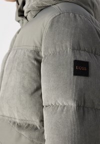 Kurtka puchowa w jasnoszarym kolorze, z fakturowaną tkaniną sztruksową na górnej części i gładką dolną częścią, z czarną naszywką z logo.
