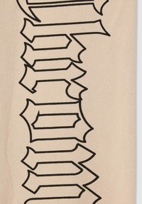 Beżowy materiał z wyraźnym czarnym graficznym tekstem, z ostrymi kształtami na końcach i teksturowaną powierzchnią, ustawiony pionowo wzdłuż materiału.