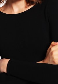 Zwarte blouse met lange mouwen en een ronde halslijn. Zacht, gestructureerd materiaal met een aansluitende pasvorm. Close-up van de armen gekruist bij de polsen.