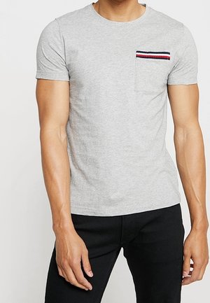Mann trägt ein eng anliegendes hellgraues Kurzarm-T-Shirt mit einer kleinen Brusttasche mit blauen, weißen und roten Streifen, kombiniert mit einer schwarzen Hose.
