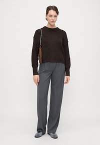 Brauner gestrickter Pullover mit Rundhalsausschnitt, kombiniert mit grauen, weiten Hosen. Das Outfit umfasst eine schwarze Tasche mit einem Quastenakzent.