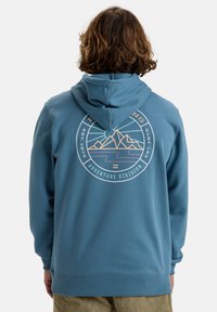 Modrá mikina s kapucí vyrobená z měkkého materiálu, která má kulatý grafický design s horami a textem "Adventure Division" v pastelových barvách.