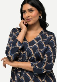 Marinblå tunika med ett abstrakt mönster i beige och ljusblått, med V-ringning och trekvartslånga ärmar, tillverkad av mjukt tyg.