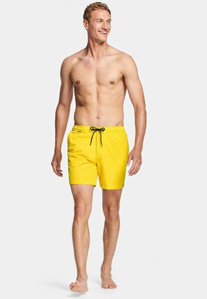 Man loopt op blote voeten, draagt felgele zwembroek met zwarte trekkoord, lacht en kijkt naar de zijkant tegen een effen achtergrond.