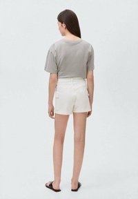 Grå kortärmad skjorta med en avslappnad passform, kombinerad med vita shorts med sidofickor. Modellen bär svarta tofflor.