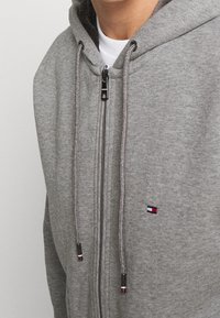 Graue Zip-Hoodie aus weichem Material. Verfügt über zweifarbige Kordelzüge, einen silbernen Reißverschluss und ein kleines Logo in Rot, Weiß und Blau auf der Brust.