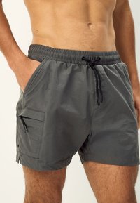 Grijze zwembroek met elastische tailleband, sluiting met trekkoord, zijzakken en een met ritssluiting afsluitbare zak op het dijbeen. Gladde textuur, knielengte.