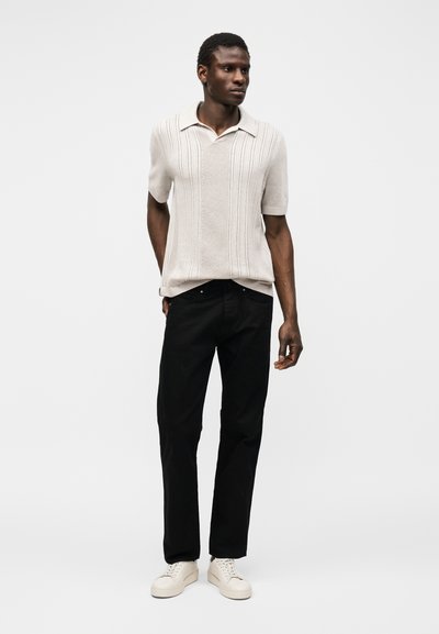 Άνδρας που φοράει μπεζ πλεκτό πόλο με κοντό μανίκι, μαύρο παντελόνι και άσπρα αθλητικά παπούτσια, στέκεται μπροστά σε απλό λευκό φόντο.