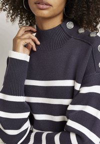 Gros plan sur une personne portant un pull à rayures bleu marine et blanc avec des boutons argentés sur l'épaule, un col montant côtelé et des poignets côtelés.