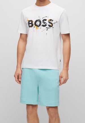 Mann trägt weißes kurzärmeliges T-Shirt mit "BOSS" in schwarzem Farbspritzer-Druck und hellblaue knielange Shorts mit Logo-Patch.