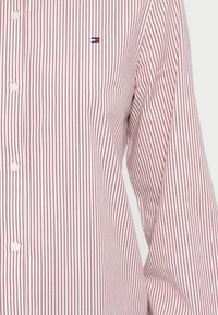 Chemise rayée rouge et blanche avec un devant boutonné, des manches longues et un petit logo brodé. Le tissu semble lisse et léger.