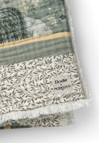 Rektangulær tæppe med en blød, plyssede underside. Har et blomstermotiv i grøn og creme, afgrænset af et bladmønster. Etikette er til stede.