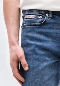 Κοντινό πλάνο του δεξιού χεριού που ακουμπά δίπλα σε σκούρο μπλε τζιν με το λογότυπο Calvin Klein στην τσέπη και μέρος ενός άσπρου πουκάμισου ορατό.