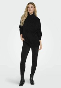 Maglione turtleneck nero, oversize, con una texture morbida, abbinato a pantaloni neri a vita alta, aderenti e stivali alla caviglia, in piedi contro uno sfondo semplice.