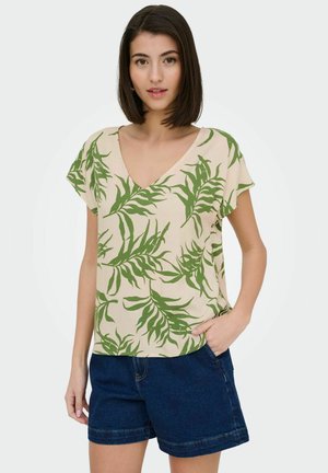 Young woman with straight dark hair wearing beige V-neck shirt with green leaf print and dark denim shorts, one hand in pocket.