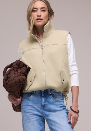 Donna bionda che indossa un gilet beige senza maniche con zip, una maglia bianca a maniche lunghe e jeans azzurri chiari, che tiene in mano sinistra una borsa marrone trapuntata.