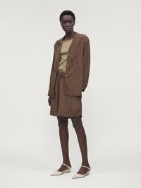 Femme portant une veste marron à motifs boutonnée et un short assorti avec un haut vert clair et des ballerines beiges pointues.