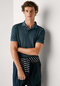 Mladý muž kučeravých vlasov v tmavomodrozelenom polokošeli a pruhovanom svetri zviazanom okolo pása, stojaci pri bielej stene.