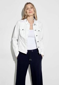 Giacca di denim bianca con chiusure a bottone e tasche sul petto, indossata sopra un top bianco e pantaloni a gamba larga blu scuro con cordoncino.