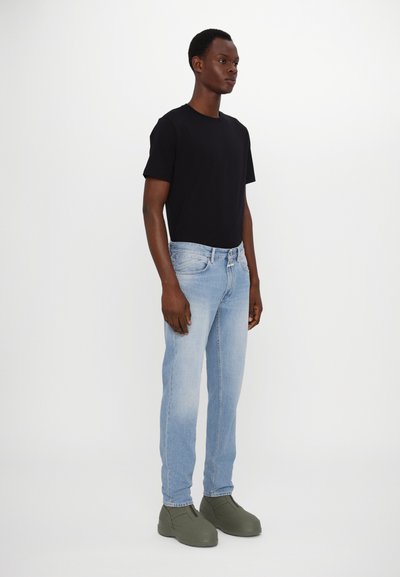 T-shirt noir à manches courtes, jeans bleu clair et chaussures sans lacets vertes. La tenue présente des lignes simples et épurées ainsi qu'une coupe décontractée.