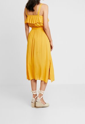 Keltainen midi-mekko, jossa on röyhelöolkaimet ja kapea vyötärö, sisältäen sivuhalkion. Yhdistetty vaaleanbeigeihin espadrille-korkokenkiin, joissa on nauhakiinnitys.