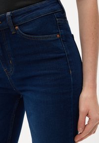 Temno modri kavbojki z oprijetim krojem, z oranžnim šivom in minimalnim dodatnim okrasjem. Podrobnosti žepa so vidne na strani.