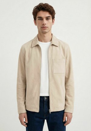 Ung mann med kort brunt hår iført en beige jakke med glidelås over en hvit skjorte og mørkeblå jeans mot en enkel lys bakgrunn.