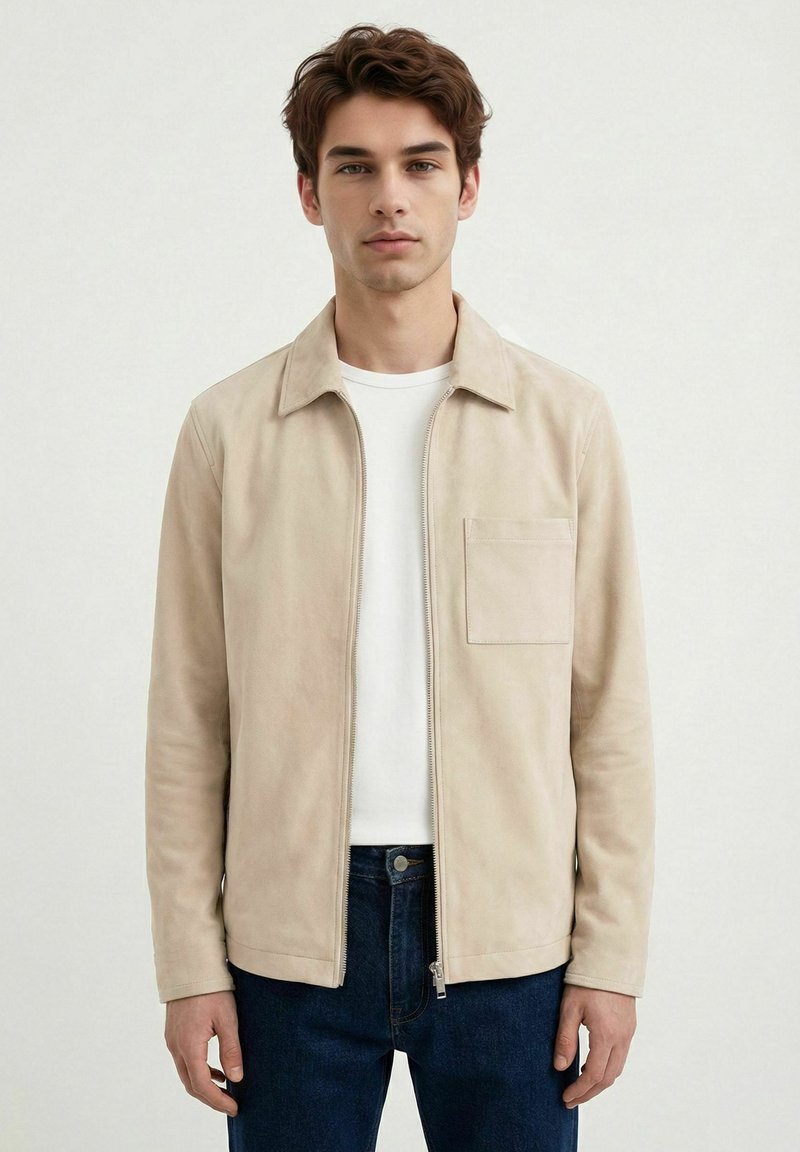 Ung mann med kort brunt hår iført en beige jakke med glidelås over en hvit skjorte og mørkeblå jeans mot en enkel lys bakgrunn.