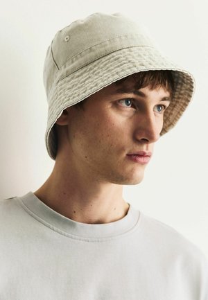 Junger Mann mit blauen Augen, der einen hellbeigen Anglerhut und ein schlichtes hellgraues Rundhalsshirt trägt und auf einem weißen Hintergrund nach rechts schaut.